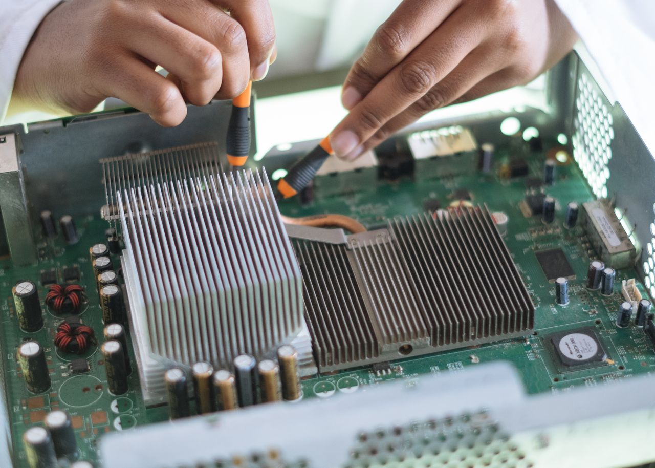 Unrecognizable ethnic engineer in white coat checking video card on computer while using ammeter in service center